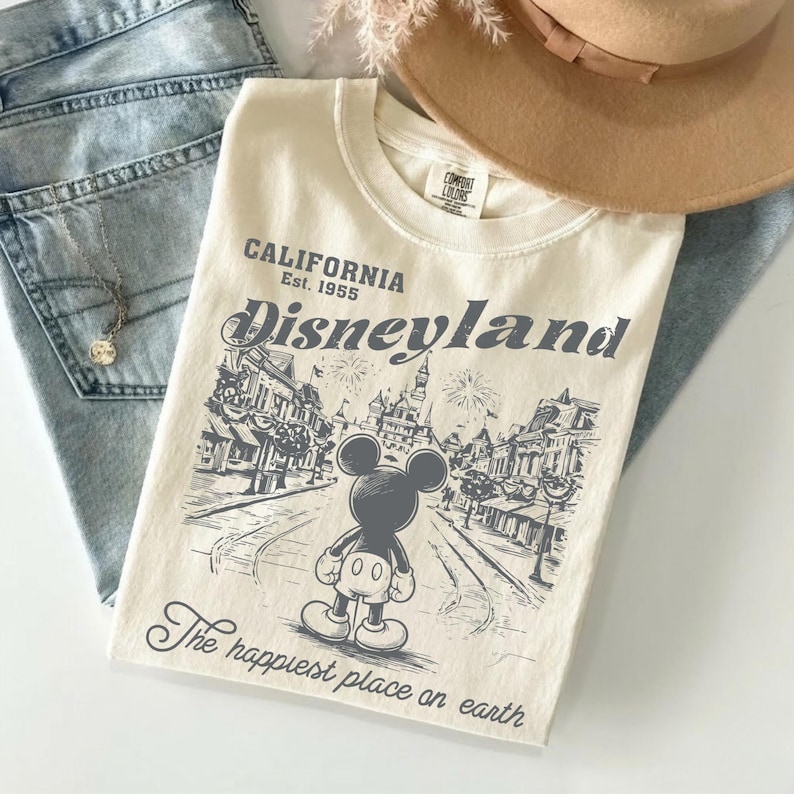 May include: Cream-colored t-shirt featuring a Disneyland graphic with Mickey Mouse. The shirt includes the text "CALIFORNIA Est. 1955" and "Disneyland" above the graphic, and "The happiest place on earth" below. The shirt is paired with blue jeans and a brown hat.