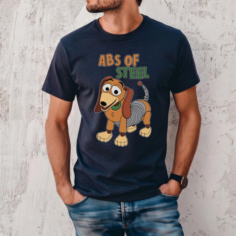 K&ouml;nnte beinhalten: Marineblaues T-Shirt mit einem Cartoon-Bild von Slinky Dog und dem Text "ABS OF STEEL" in Gr&uuml;n und Orange. Der Hund hat braune Ohren, einen gr&uuml;nen Kragen und einen silbernen Federk&ouml;rper. Das T-Shirt wird von einer Person getragen.