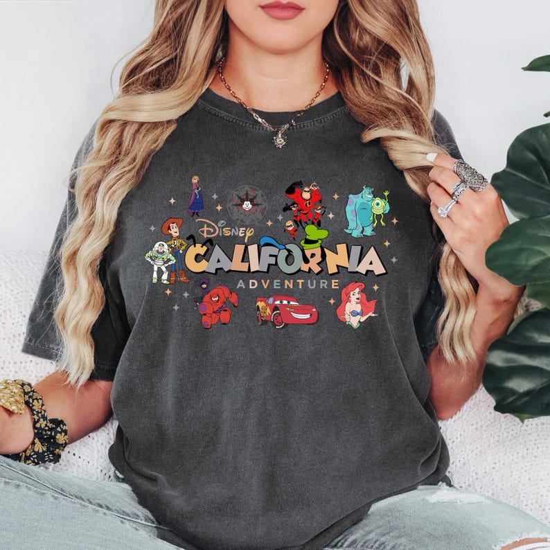 K&ouml;nnte beinhalten: Ein anthrazitfarbenes T-Shirt mit einem Grafikdruck und dem Text Disney CALIFORNIA ADVENTURE. Das Design enth&auml;lt farbenfrohe Illustrationen verschiedener Zeichentrickfiguren wie Woody, Buzz Lightyear, Baymax, Lightning McQueen, Ariel und anderen.