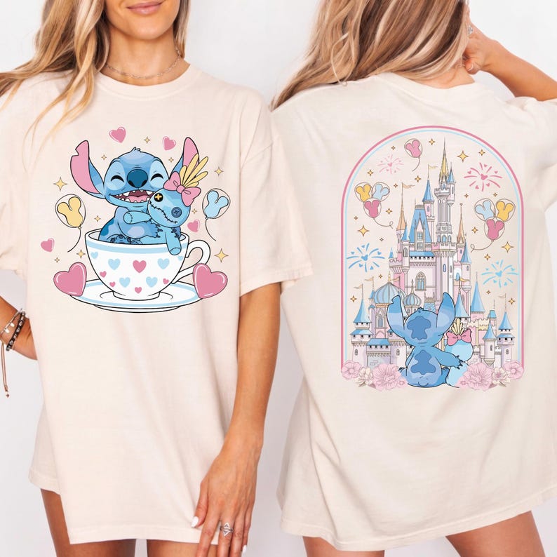 May include: Cream-colored t-shirt featuring a blue cartoon character embracing another in a teacup with hearts. The back of the shirt displays a castle scene with the same character. Pink and yellow accents complete the design.