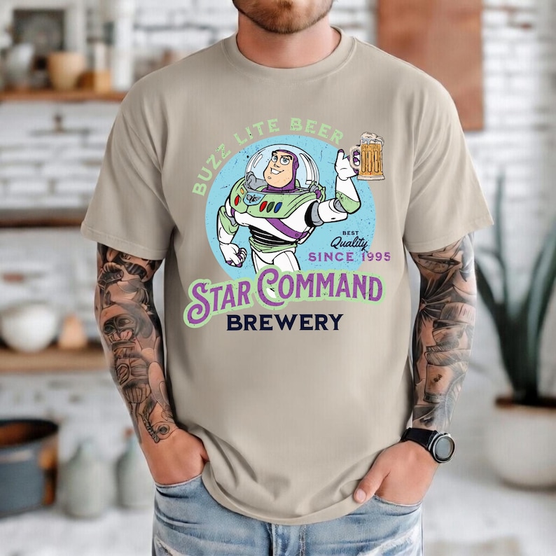 May include: A beige short-sleeve t-shirt featuring a graphic of a space ranger character holding a beer mug. The text reads BUZZ LITE BEER, BEST QUALITY SINCE 1995, STAR COMMAND BREWERY. The design is set against a light blue circular background.