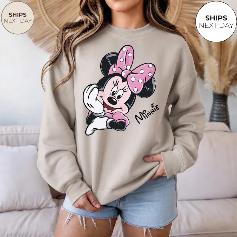 May include: Beige sweatshirt featuring a Minnie Mouse graphic. The graphic shows Minnie Mouse with a pink polka dot bow, black ears, and the name "Minnie" in script. The sweatshirt is paired with denim shorts.