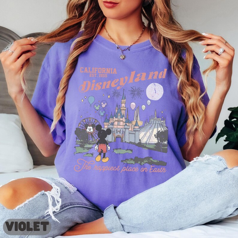 May include: Violet-colored t-shirt featuring a Disneyland graphic. The design includes the text "California Est. 1955 Disneyland" with an illustration of the castle, fireworks, and Mickey Mouse. The phrase "The happiest place on Earth" is also included.