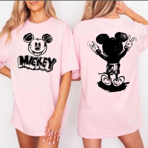 May include: Light pink t-shirts featuring a black and white Mickey Mouse design. The front of the shirt displays Mickey's face with the word "Mickey" below. The back of the shirt shows Mickey with his arms up in a rock and roll pose.