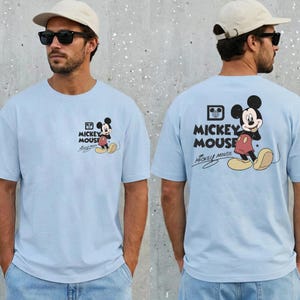 May include: Light blue t-shirt featuring Mickey Mouse. The front of the shirt has a small graphic of Mickey Mouse, while the back has a larger design with the words "Mickey Mouse".
