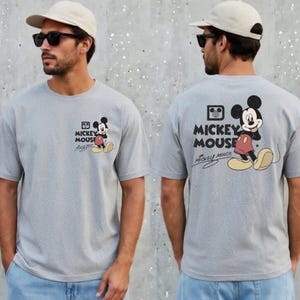 May include: A light gray t-shirt featuring Mickey Mouse. The front of the shirt has a small graphic of Mickey Mouse with the words "Mickey Mouse" and a signature. The back of the shirt has a larger Mickey Mouse graphic and the words "Mickey Mouse".