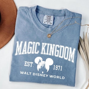 May include: A blue t-shirt with the words "MAGIC KINGDOM" in white block letters. Below the text is a white graphic of Mickey Mouse ears and a castle, with the text "EST. 1971 WALT DISNEY WORLD". A gold necklace rests on the shirt.