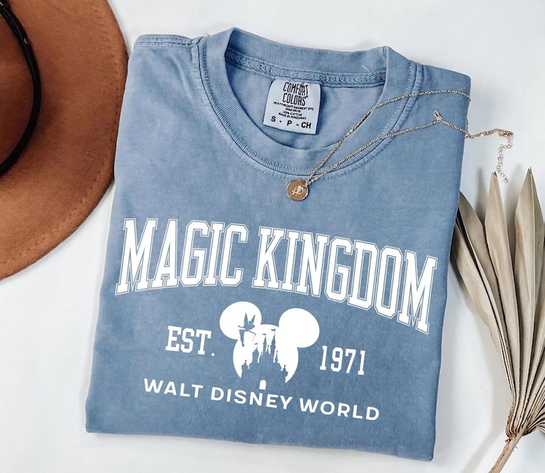 May include: A blue t-shirt with the words "MAGIC KINGDOM" in white block letters. Below the text is a white graphic of Mickey Mouse ears and a castle, with the text "EST. 1971 WALT DISNEY WORLD". A gold necklace rests on the shirt.