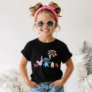 Puede incluir: Una camiseta negra con un gr&aacute;fico colorido de un personaje de dibujos animados sosteniendo globos y conos de helado. El gr&aacute;fico incluye el personaje Stitch de Lilo & Stitch. La camiseta la lleva una ni&ntilde;a con gafas de sol y una diadema rosa.