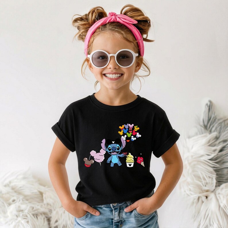 Puede incluir: Una camiseta negra con un gr&aacute;fico colorido de un personaje de dibujos animados sosteniendo globos y conos de helado. El gr&aacute;fico incluye el personaje Stitch de Lilo & Stitch. La camiseta la lleva una ni&ntilde;a con gafas de sol y una diadema rosa.