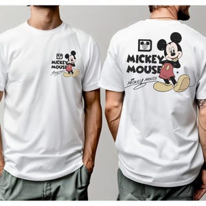 May include: White t-shirt featuring a Mickey Mouse graphic on the front and back. The front graphic is smaller, while the back graphic is larger. The design includes Mickey Mouse in his classic outfit, with the words "Mickey Mouse" and "Mickey Mouse" in a stylized font.