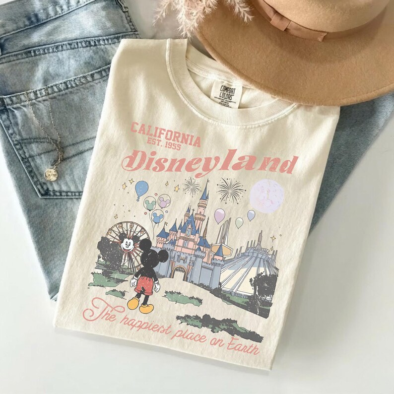 May include: Cream-colored t-shirt with a Disneyland graphic. The design features Mickey Mouse, a castle, fireworks, and the text "California Disneyland Est. 1955" and "The happiest place on Earth". The shirt is paired with jeans and a hat.