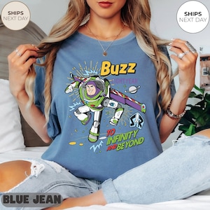 K&ouml;nnte beinhalten: Blaues T-Shirt mit einem Buzz Lightyear-Motiv, mit dem Text "Buzz Lightyear" und "To Infinity and Beyond". Das Design ist in Gr&uuml;n, Lila, Wei&szlig; und Gelb gehalten. Das T-Shirt hat eine lockere Passform.