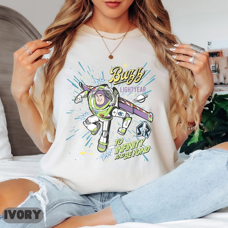 May include: Ivory-colored t-shirt featuring a graphic of Buzz Lightyear from Toy Story. The design includes the text "Buzz Lightyear" and "To Infinity and Beyond" in a colorful, cartoon style. The shirt is a crew neck and has short sleeves.