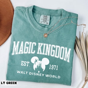 May include: A light green t-shirt with the words "MAGIC KINGDOM" in white block letters. Below the text is "EST. 1971 WALT DISNEY WORLD" in white. The shirt has a crew neck and short sleeves. A gold necklace with a pendant rests on the shirt.