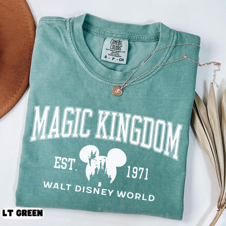 May include: A light green t-shirt with the words "MAGIC KINGDOM" in white block letters. Below the text is "EST. 1971 WALT DISNEY WORLD" in white. The shirt has a crew neck and short sleeves. A gold necklace with a pendant rests on the shirt.