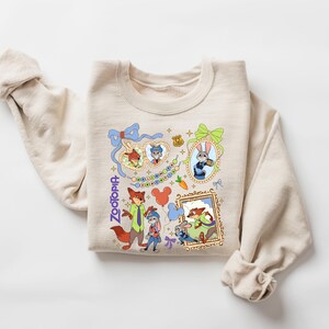 Może przedstawiać: Beżowa bluza z kolorowymi ilustracjami z kresk&oacute;wki z filmu Zwierzogr&oacute;d. Projekt zawiera postacie takie jak Nick Wilde i Judy Hopps, a także słowo "Zootopia" w niebieskim skrypcie. Bluza ma długie rękawy.