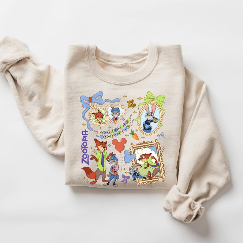 Może przedstawiać: Beżowa bluza z kolorowymi ilustracjami z kresk&oacute;wki z filmu Zwierzogr&oacute;d. Projekt zawiera postacie takie jak Nick Wilde i Judy Hopps, a także słowo "Zootopia" w niebieskim skrypcie. Bluza ma długie rękawy.