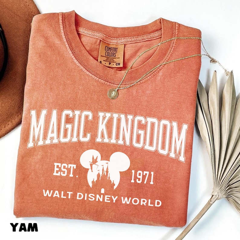 May include: A coral-colored crew neck t-shirt with the words "MAGIC KINGDOM" in white block letters. Below the text is "EST. 1971" and "WALT DISNEY WORLD" with a Mickey Mouse silhouette. A gold necklace with a pendant rests on the shirt.
