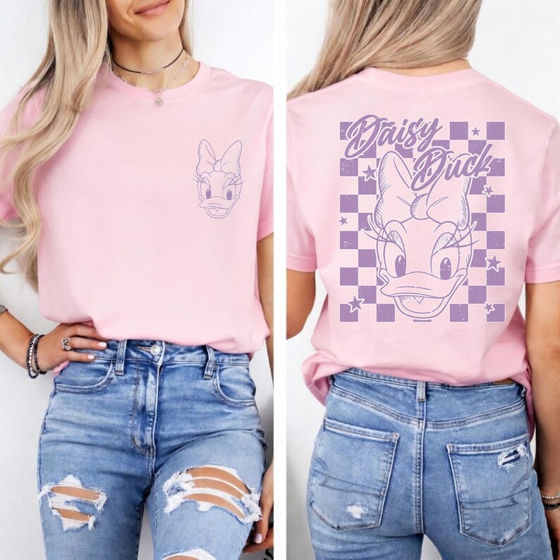 May include: A light pink t-shirt featuring a purple Daisy Duck graphic on the front and back. The front has a small graphic, while the back displays a larger design with the text "Daisy Duck" and a checkered pattern.