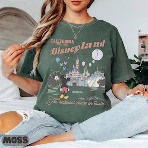May include: Green t-shirt with a Disneyland graphic. The design features the text "California Est. 1955 Disneyland" above an illustration of the castle, Mickey Mouse, and a Ferris wheel. The text "The happiest place on Earth" is below the graphic.