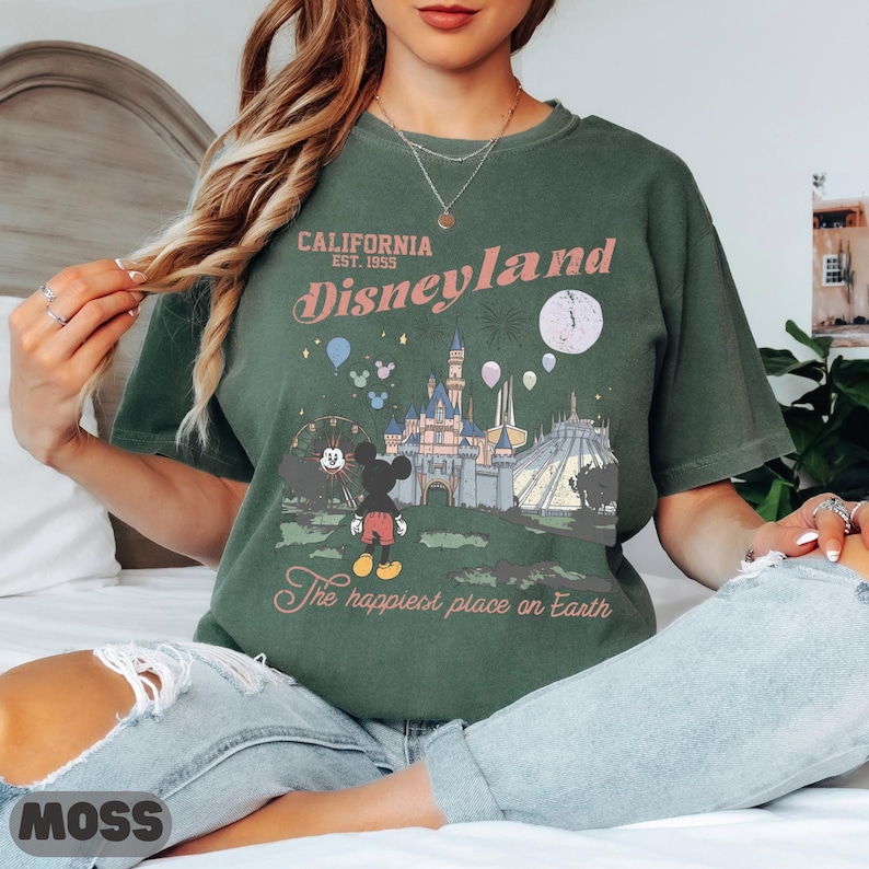 May include: Green t-shirt with a Disneyland graphic. The design features the text "California Est. 1955 Disneyland" above an illustration of the castle, Mickey Mouse, and a Ferris wheel. The text "The happiest place on Earth" is below the graphic.
