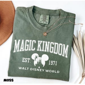 May include: An olive green t-shirt with the words "MAGIC KINGDOM" in white block letters. Below the text is "EST. 1971" and "WALT DISNEY WORLD" with a Mickey Mouse silhouette. A gold necklace rests on the shirt.