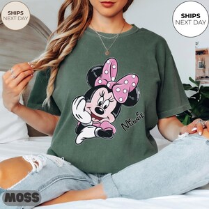 May include: Green t-shirt featuring a Minnie Mouse graphic. The cartoon character is depicted with a pink bow with white polka dots, a pink dress, and the name "Minnie" in script. The shirt is a relaxed fit.
