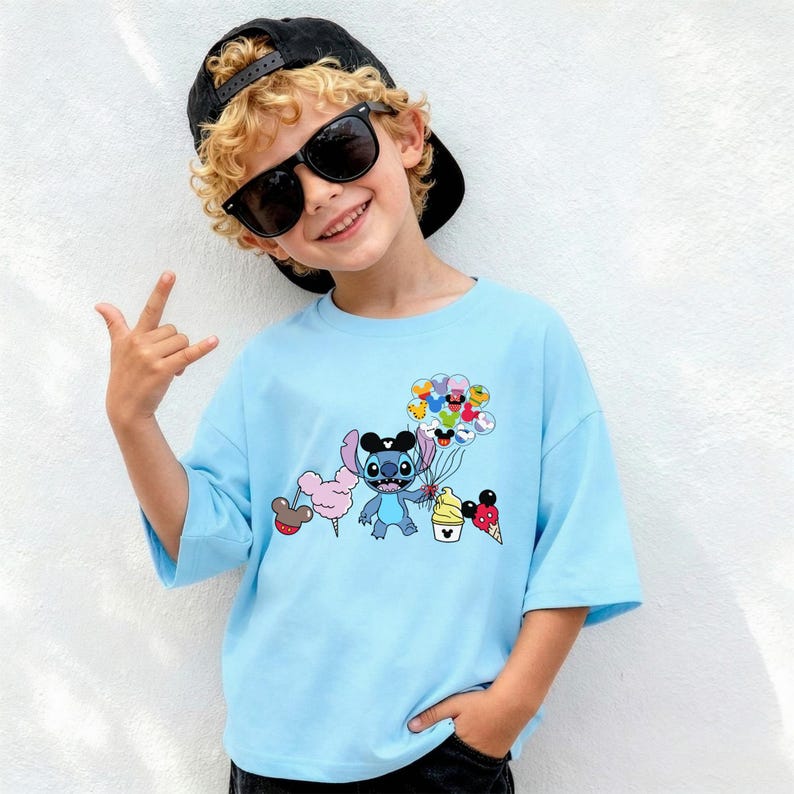Puede incluir: Camiseta azul claro con un personaje de dibujos animados sosteniendo globos, helado y algod&oacute;n de az&uacute;car. La camiseta tiene mangas cortas y un corte holgado. La persona lleva una gorra negra y gafas de sol.