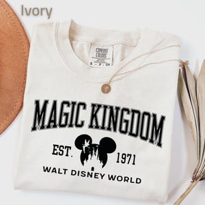 May include: Ivory-colored t-shirt with the words "MAGIC KINGDOM" in black block letters. Below the text is a silhouette of Mickey Mouse ears and a castle, with "EST. 1971" and "WALT DISNEY WORLD" printed below. A gold necklace is draped across the shirt.