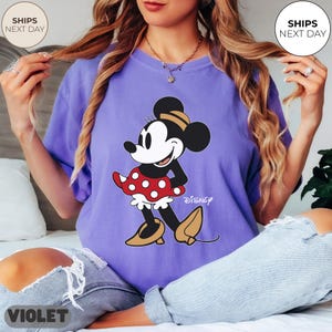 Puede incluir: Camiseta violeta con un gr&aacute;fico de Minnie Mouse. El gr&aacute;fico muestra a Minnie Mouse con una falda roja de lunares, una parte superior negra y zapatos amarillos. La palabra "Disney" est&aacute; impresa debajo. La camiseta tiene un corte holgado.