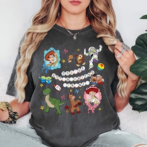 May include: Dark grey t-shirt with a colourful cartoon design featuring characters from a popular animated film, including Woody and Buzz Lightyear. The text "YOU'VE GOT A FRIEND IN ME" is also displayed.