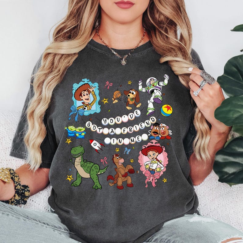 May include: Dark grey t-shirt with a colourful cartoon design featuring characters from a popular animated film, including Woody and Buzz Lightyear. The text "YOU'VE GOT A FRIEND IN ME" is also displayed.