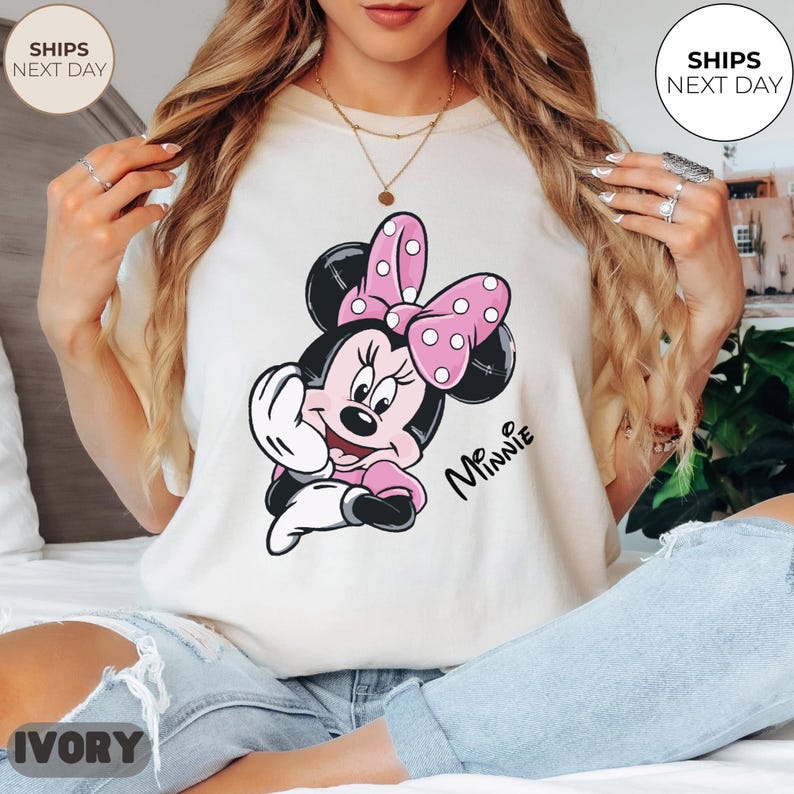 May include: Ivory-colored t-shirt featuring a Minnie Mouse graphic. The cartoon character is depicted in black, pink, and white, with a pink polka-dot bow. The word "Minnie" is written in cursive. The shirt is a casual wear item.