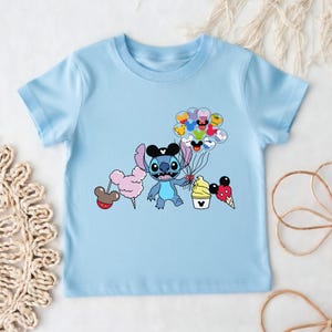 Puede incluir: Camiseta azul claro con un personaje de dibujos animados que sostiene globos y golosinas. El personaje azul tiene ojos grandes y orejas negras. La camiseta tiene un gr&aacute;fico de dibujos animados del personaje, algod&oacute;n de az&uacute;car, helado y globos.