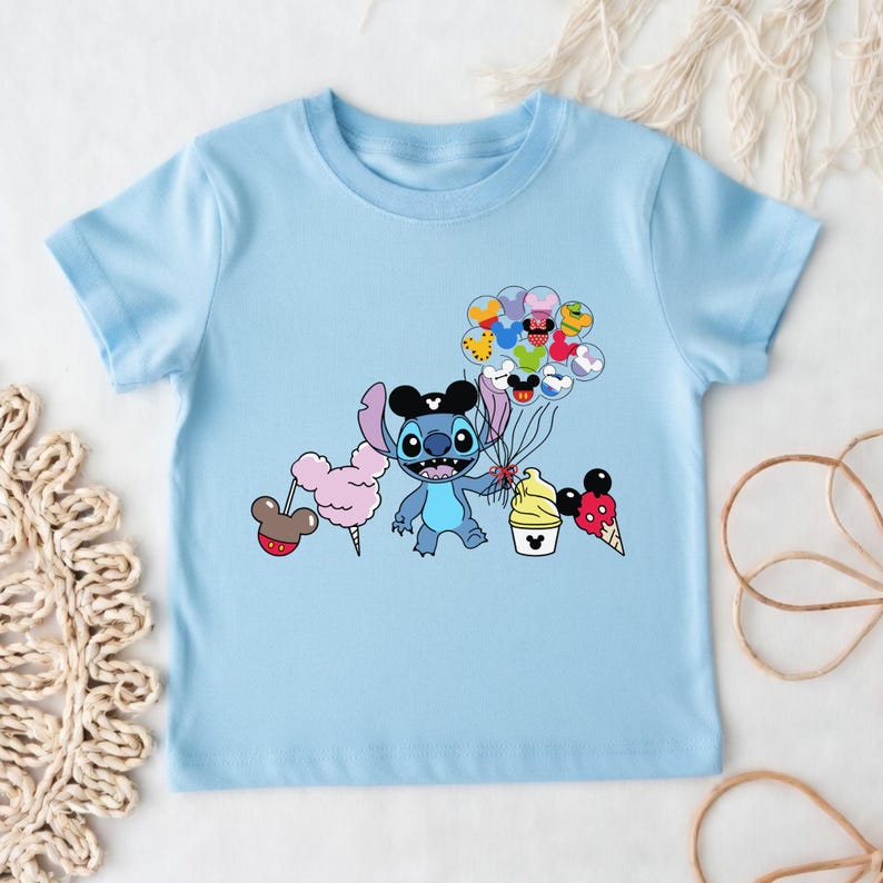 Puede incluir: Camiseta azul claro con un personaje de dibujos animados que sostiene globos y golosinas. El personaje azul tiene ojos grandes y orejas negras. La camiseta tiene un gr&aacute;fico de dibujos animados del personaje, algod&oacute;n de az&uacute;car, helado y globos.