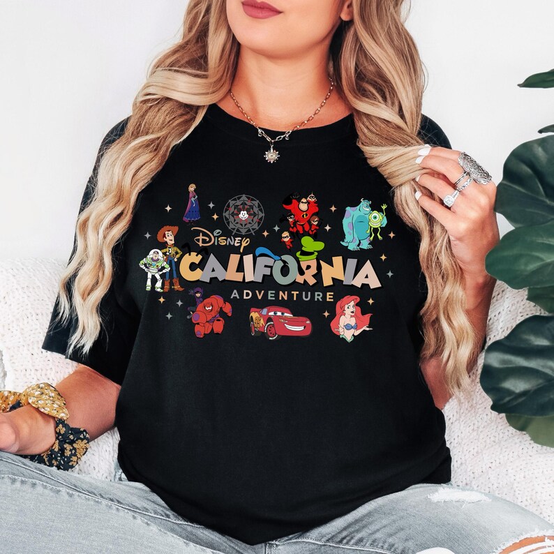 K&ouml;nnte beinhalten: Ein schwarzes T-Shirt mit einem Grafikdruck und dem Text Disney California Adventure. Das Design enth&auml;lt farbenfrohe Illustrationen verschiedener Zeichentrickfiguren wie Woody, Buzz Lightyear, Baymax, Lightning McQueen und Ariel.