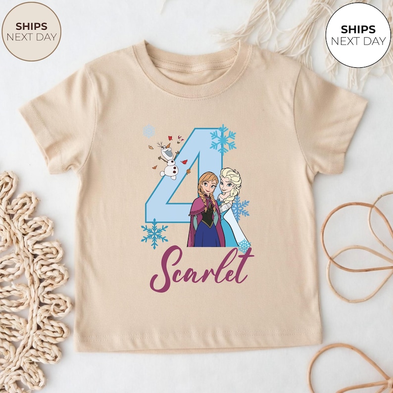 May include: A beige short-sleeved t-shirt with a blue number 4, snowflakes, and cartoon characters Anna and Elsa. The name "Scarlet" is written in a cursive font below the characters. The shirt is designed for a child.
