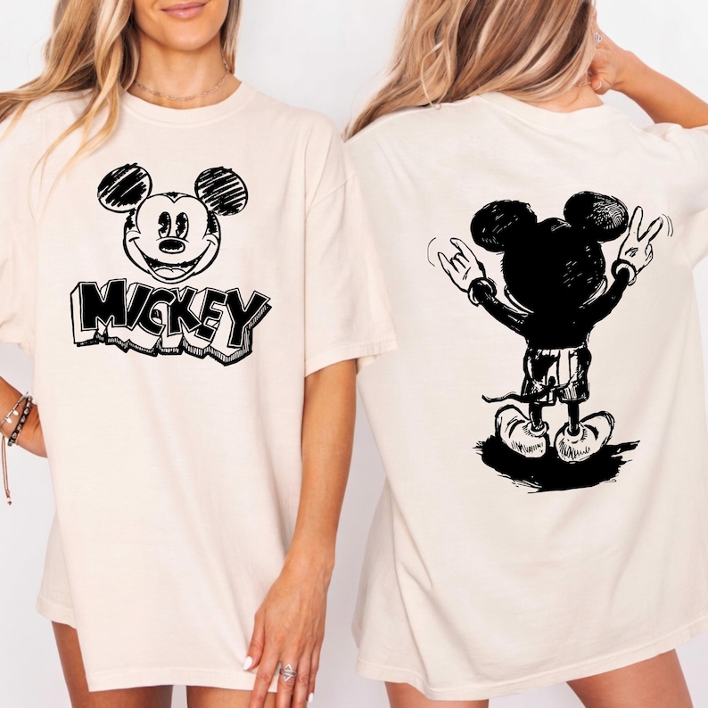 May include: Cream-colored t-shirt featuring a black and white Mickey Mouse graphic. The front of the shirt displays Mickey's face with the word "Mickey" below. The back shows Mickey with his arms raised in a rock-and-roll gesture.