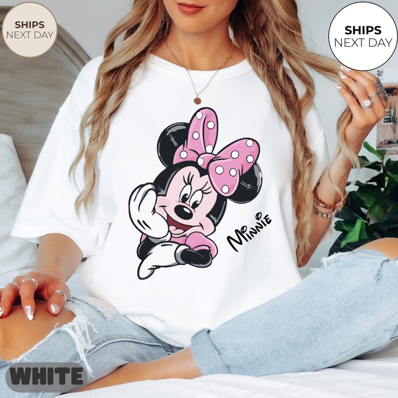 May include: White t-shirt featuring a Minnie Mouse graphic. The cartoon character is depicted in pink, black, and white, with a pink bow with white polka dots. The word "Minnie" is written in black script.