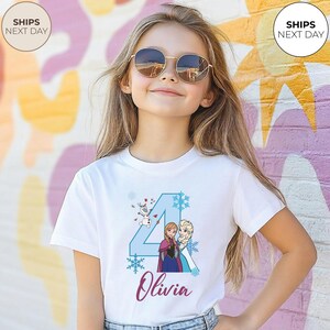 May include: A white t-shirt with a blue number 4, snowflakes, and characters Anna, Elsa, and Olaf from Frozen. The name "Olivia" is written in cursive below the graphic. The shirt is for a child.