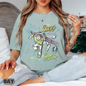 Może przedstawiać: Jasnozielona koszulka z grafiką Buzza Astrala z Toy Story. Projekt zawiera tekst "Buzz Lightyear" i "To Infinity and Beyond" w stylu kresk&oacute;wkowym. Koszulka jest wygodnym, codziennym ubraniem.
