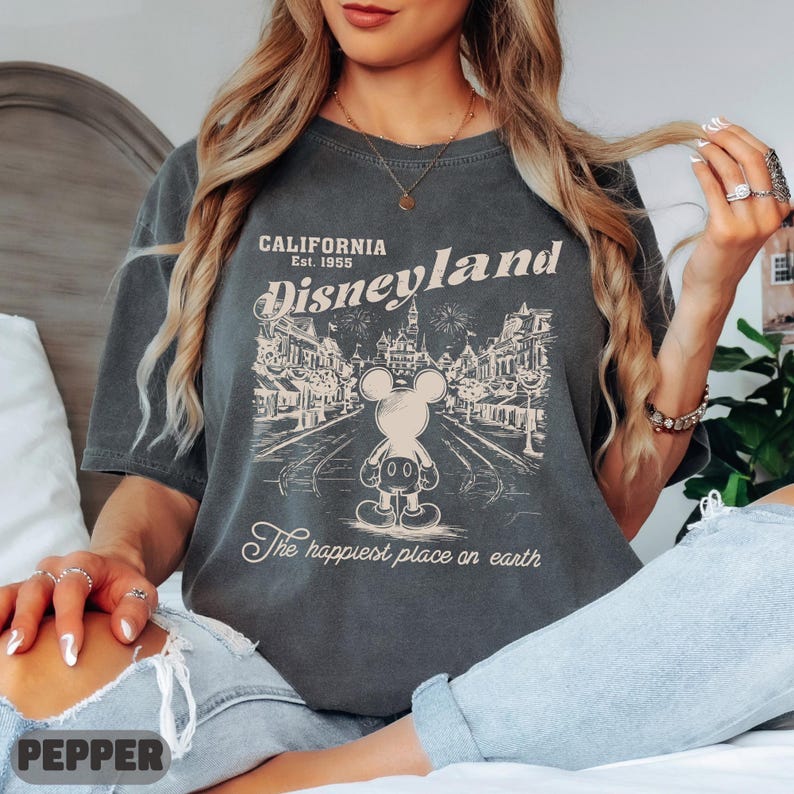 May include: Dark gray t-shirt featuring a vintage Disneyland design. The graphic includes Mickey Mouse, the words "California Disneyland Est. 1955," and "The happiest place on earth." The shirt has a classic, slightly faded look.