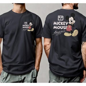 May include: Dark gray t-shirt featuring Mickey Mouse. The front of the shirt has a small Mickey Mouse graphic with the words "Mickey Mouse". The back of the shirt has a larger Mickey Mouse graphic with the words "Mickey Mouse".