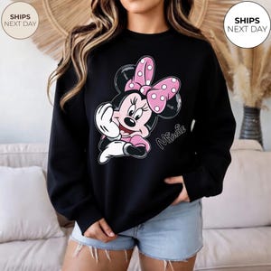 May include: Black sweatshirt featuring a Minnie Mouse graphic. The graphic shows Minnie Mouse in pink and white, with a pink bow with white polka dots. The word "Minnie" is written in cursive. The sweatshirt is a crew neck.
