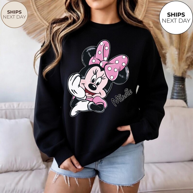 May include: Black sweatshirt featuring a Minnie Mouse graphic. The graphic shows Minnie Mouse in pink and white, with a pink bow with white polka dots. The word "Minnie" is written in cursive. The sweatshirt is a crew neck.