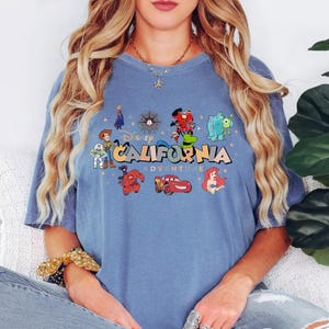 K&ouml;nnte beinhalten: Ein blaues Kurzarm-T-Shirt mit einem Grafikdruck und dem Text Disney CALIFORNIA ADVENTURE. Das Design zeigt verschiedene Zeichentrickfiguren wie Woody, Buzz Lightyear, Baymax, Lightning McQueen und Ariel, umgeben von kleinen Sternen.