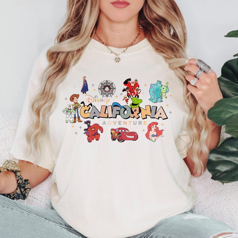 K&ouml;nnte beinhalten: Ein cremefarbenes T-Shirt mit einem bunten Grafikdruck und dem Text Disney California Adventure. Das Design zeigt verschiedene animierte Figuren wie Woody, Buzz Lightyear, Baymax, Lightning McQueen, Ariel und weitere.