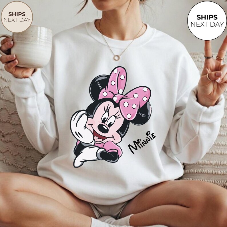 May include: White crewneck sweatshirt featuring a Minnie Mouse graphic. The graphic shows Minnie Mouse in a pink dress and bow with white polka dots. The word "Minnie" is written in a script font.