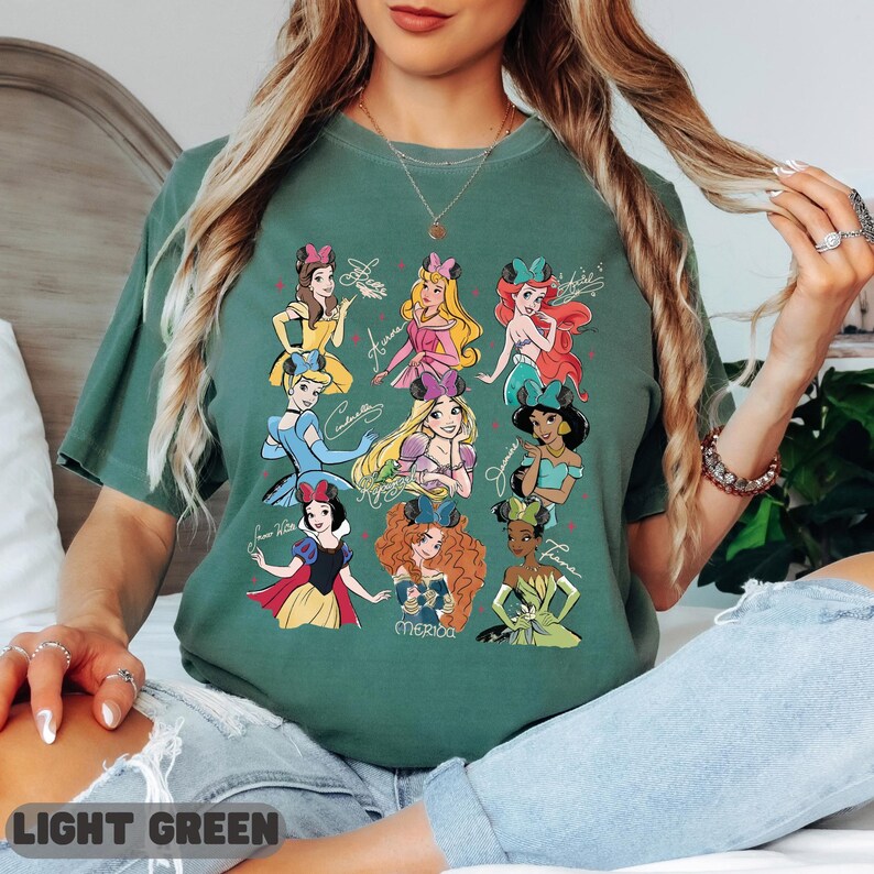 Op de afbeelding: Lichtgroen t-shirt met illustraties van Disney-prinsessen en hun namen in cursief. De prinsessen zijn in verschillende kleuren, waaronder geel, roze, blauw, rood en groen. De tekst "LIGHT GREEN" staat onderaan het shirt.
